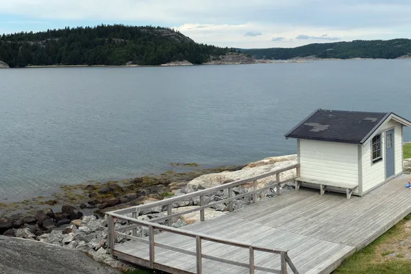 Liten vit stuga på brygga vid fjord med skogbevuxna kullar